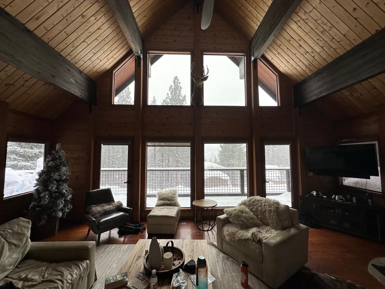 A cozy cabin living room, with wood panel walls, lots of fuzzy, furry pillows, and a high steeply pitched roof. A fireplace is off to the right, and a large couch occupies the middle of the room. The windows fill the wall, floor to ceiling. Outside, you can see many tall pine trees and snow falling.