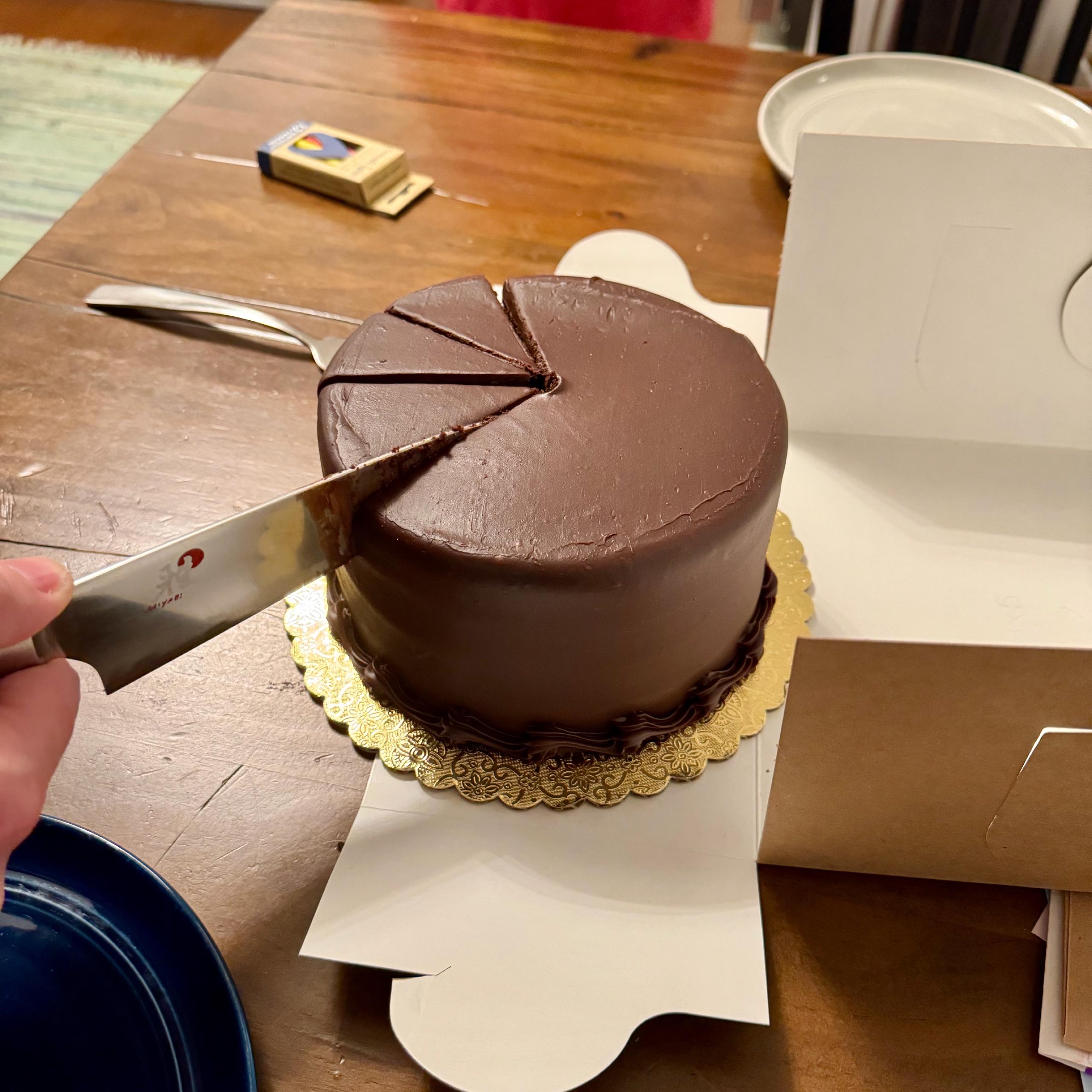 A cylidrical chocolate cake covered in smooth chocolate icing. Small flourishes adorn the bottom. Tess' hand holds a knife, part way through making a cut. Several more cuts mark out three pieces of cake ready to be served.
