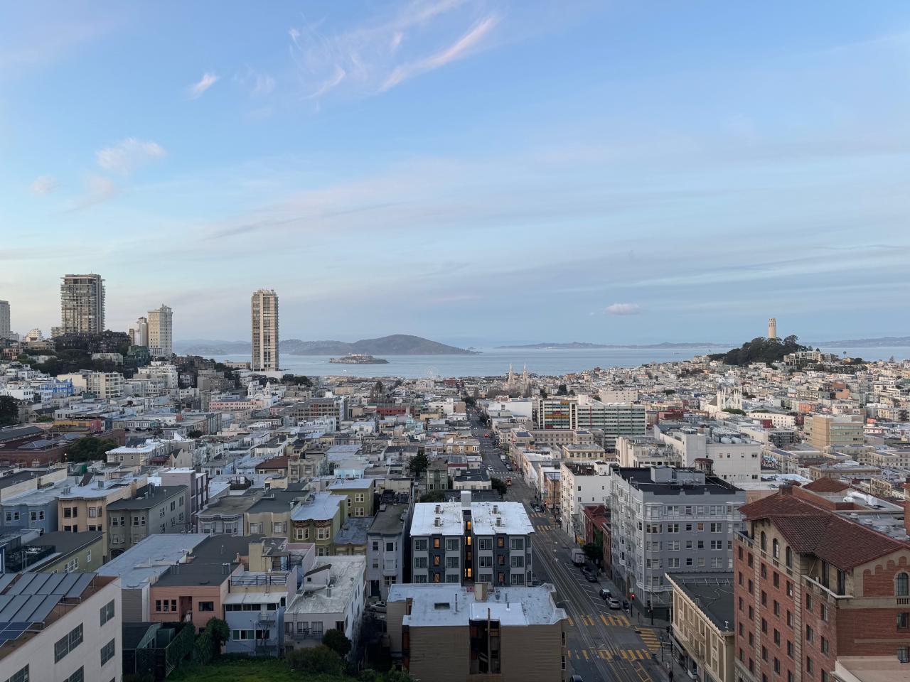  A sweeping view of San Francisco from Nob Hill on a clear day with high wispy clouds. In the center distance, Alcatraz Island rises out of the water. To the right, Coit Tower stands atop Telegraph Hill. To the left is Russian Hill, with myriad tall apartment buildings. 
