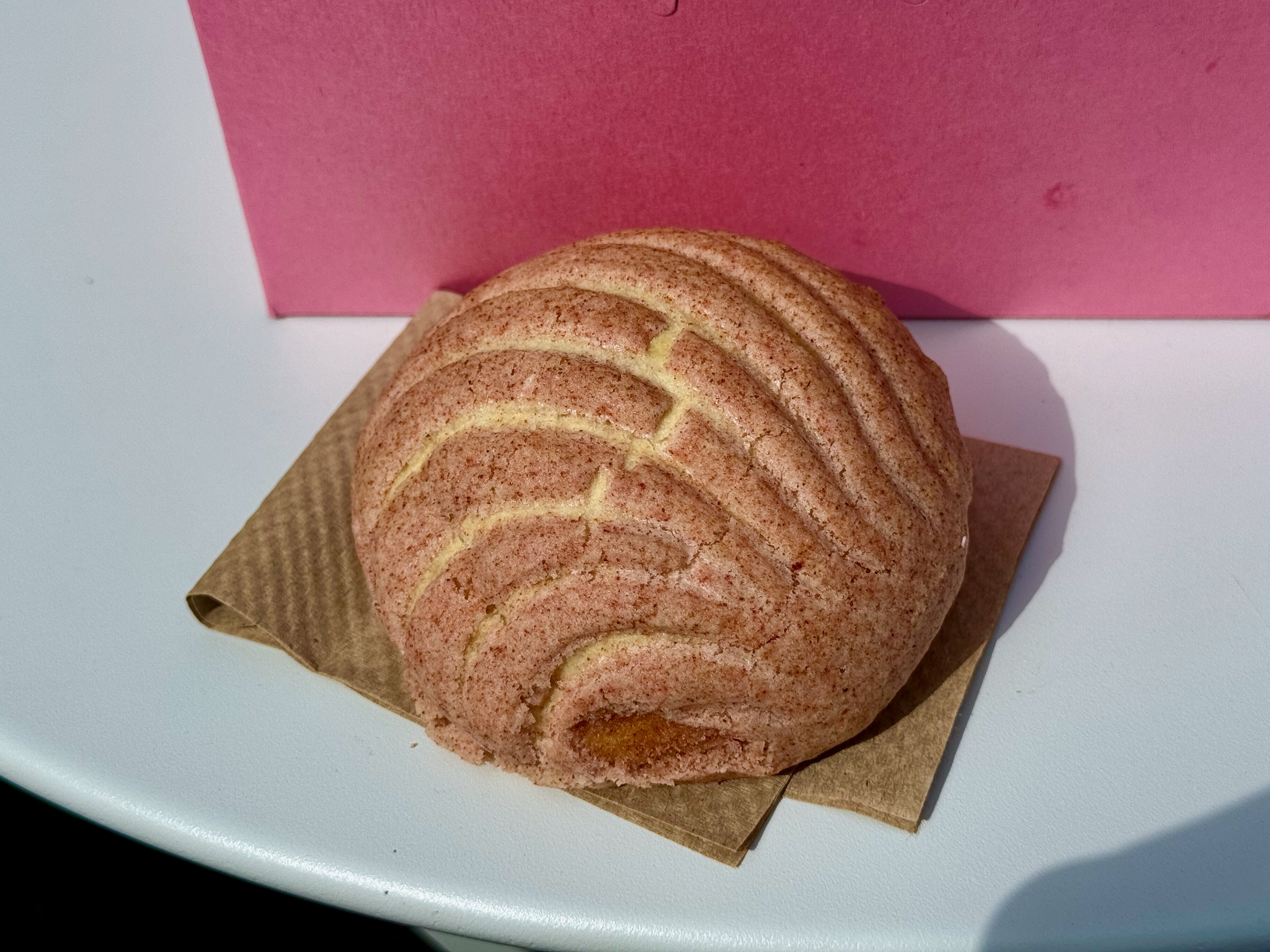 A small round bun with a pinkish topping sittin on a napkin in front of a traditional cardboard donut box.
