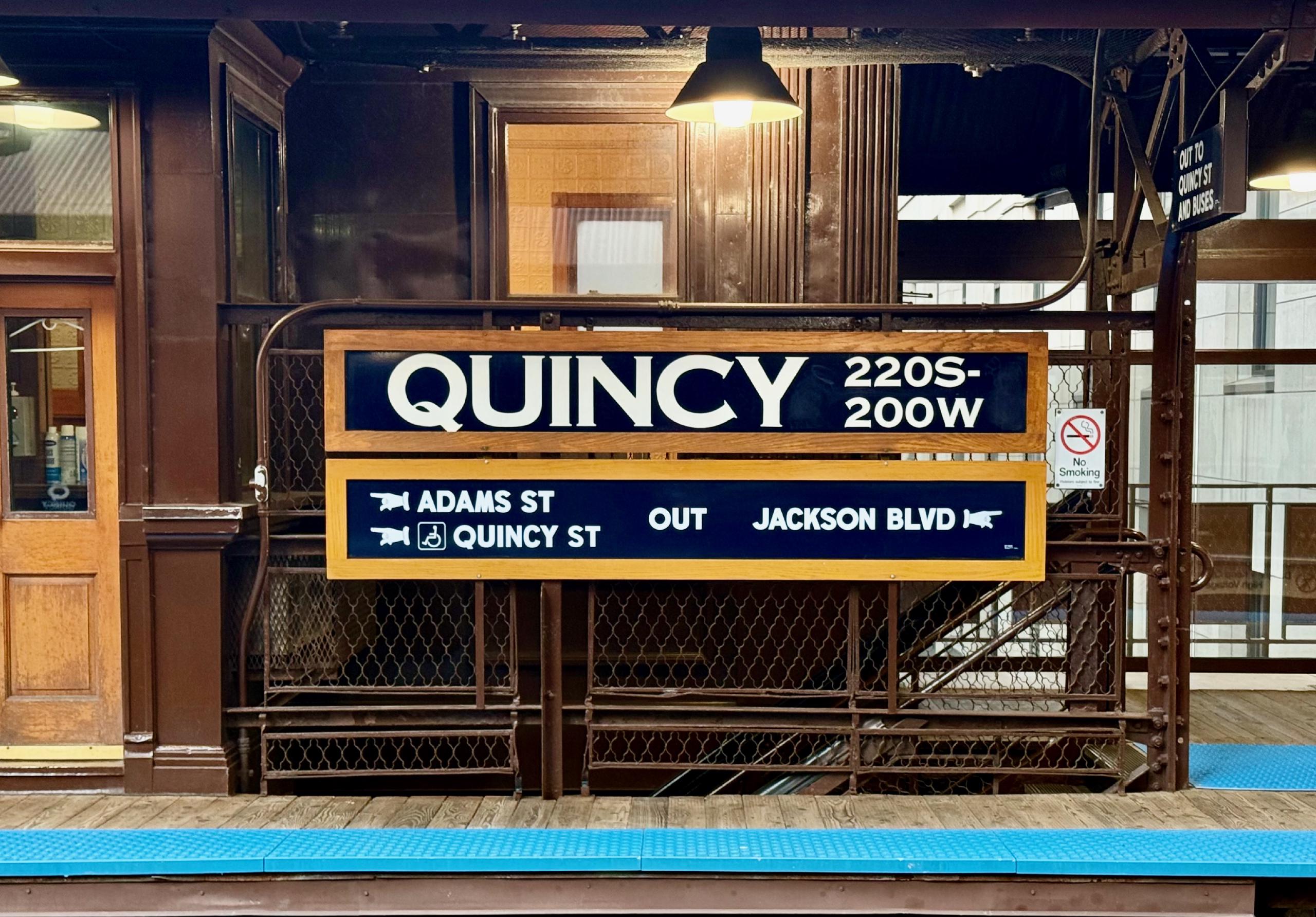 Wood plank platforms, Copperplate lettering, and wingding-style hands to direct you where you need to go.
A large station sign, framed with a simple wood frame, hanging on the railing around a station platform stairway. It declares the station's name, Quincy, and points to exits to the left and right.