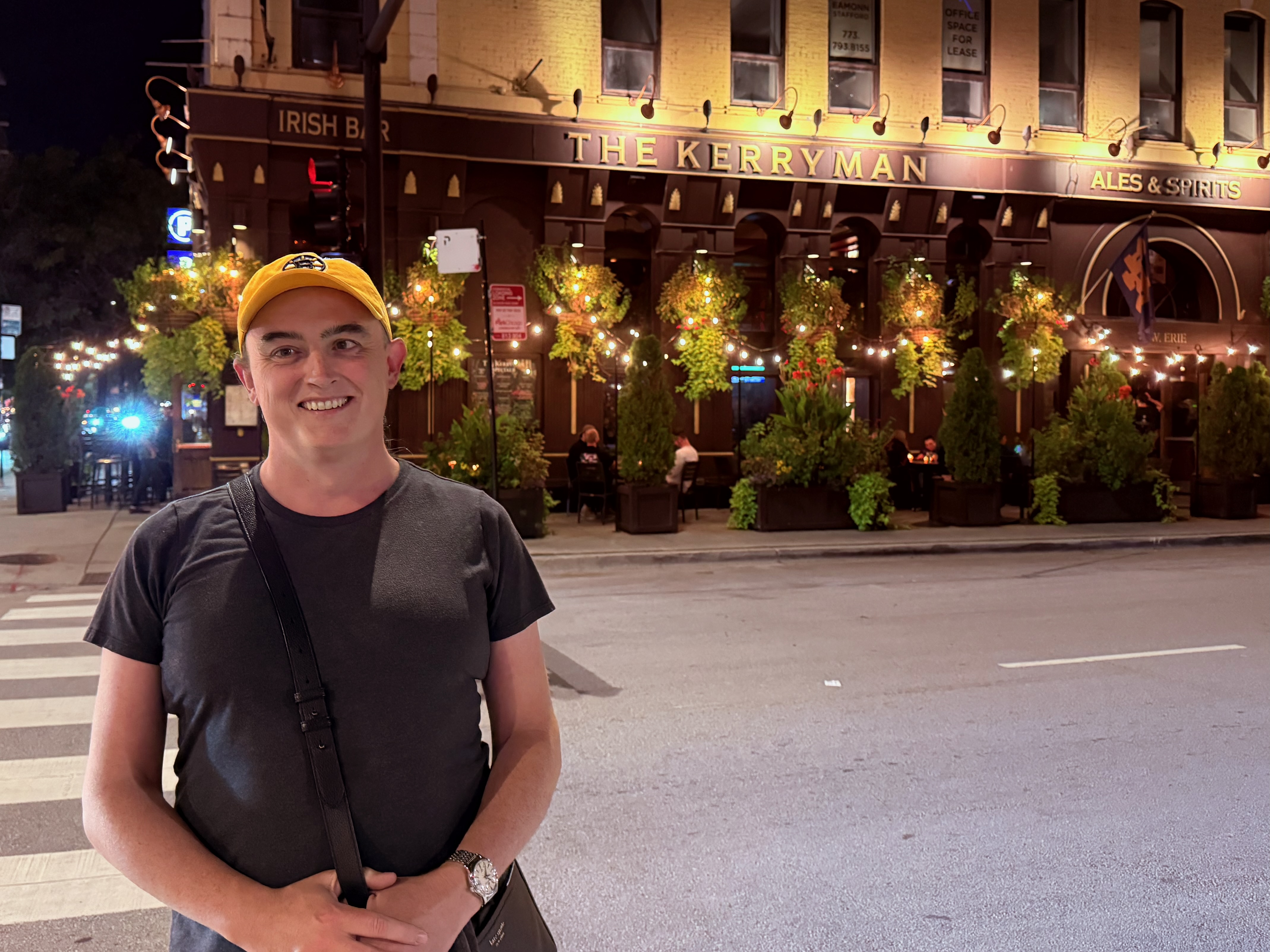 Tess, on the left, stands outside a large pub on a stree corner. The pub has outdoor seating.
