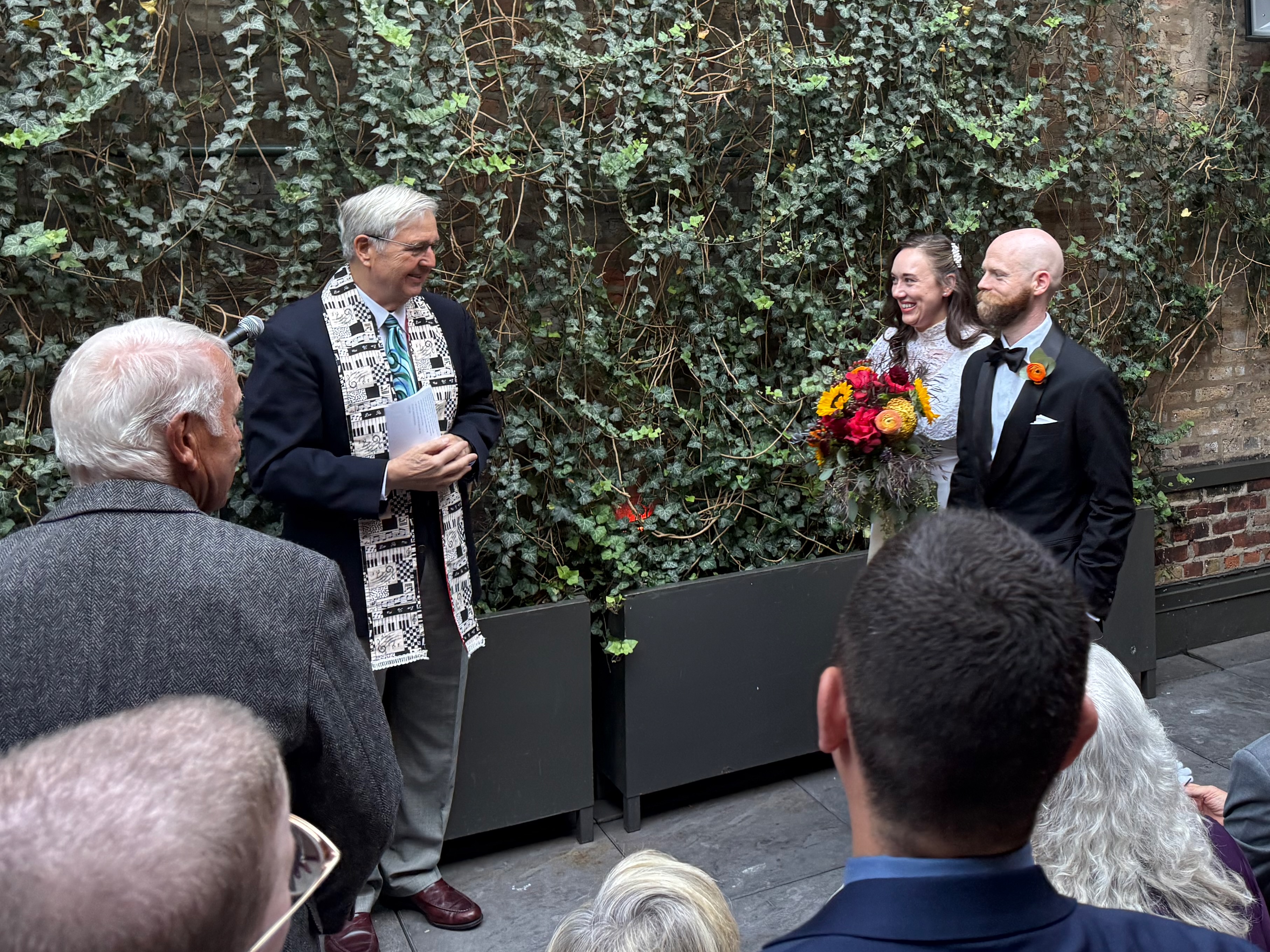 To the left, a man wearing a suit and white pastoral stole stands near a mic. To the right, a woman in a white dress, and a man in a tuxedo walk into the frame. Everyone is smiling.
