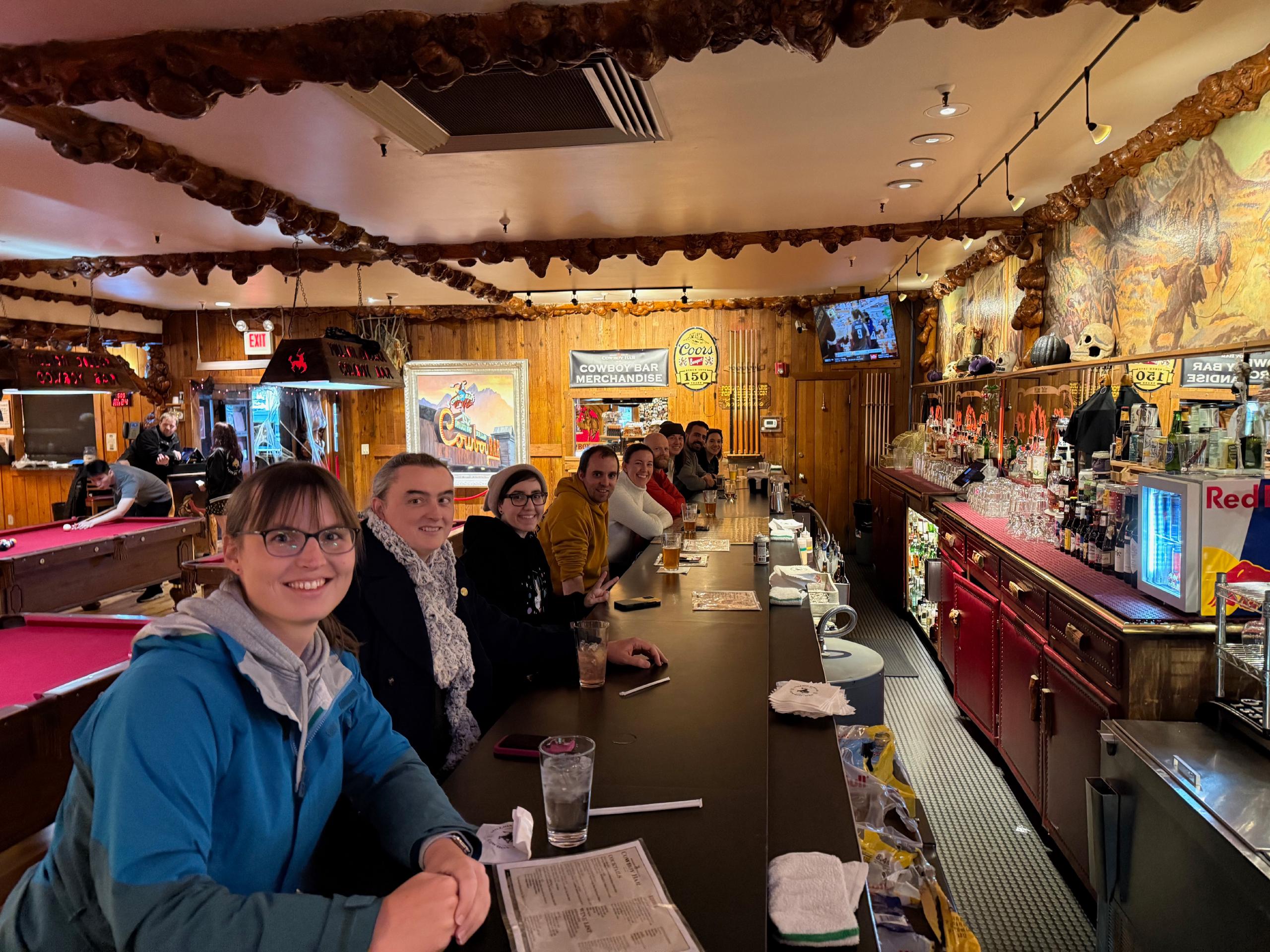 Nine people sitting along a bar, smiling at the camera. The restaurant is full of old-timey western decorations. Behind the group is a set of pool tables with red felt.
