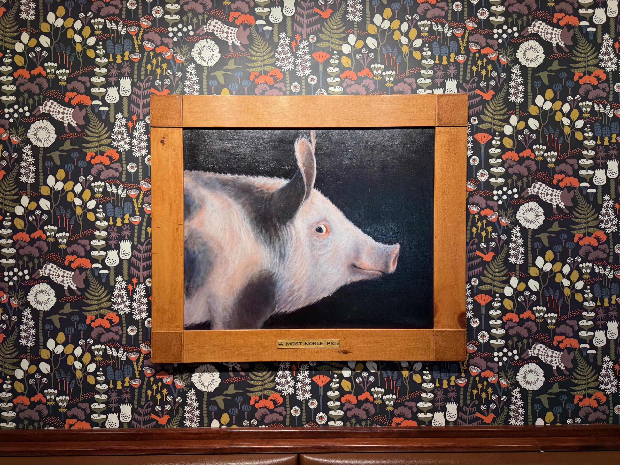 A framed painting of a pig on a dark background. The pig looks sideways at the viewer. The painting has a small caption plate saying "A Most Noble Pig". The painting is mounted on a wall covered in a busy wallpaper showing leaves, mushrooms, flowers, and pigs.
