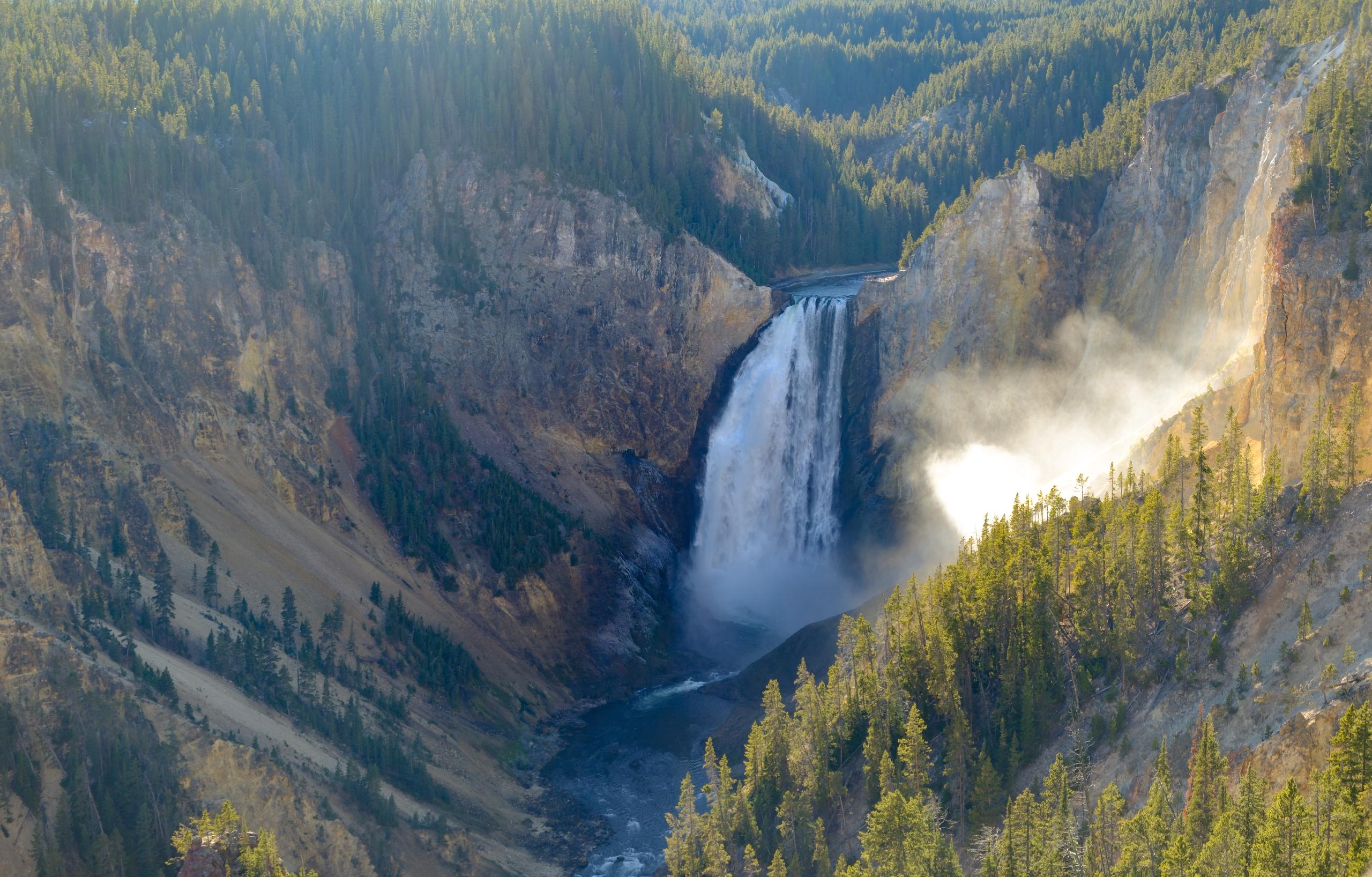 A V-shaped canyon of solid yellow stone. A large waterfall pours water into a river running along the bottom of the canyon. Trees cover the canyon walls.
