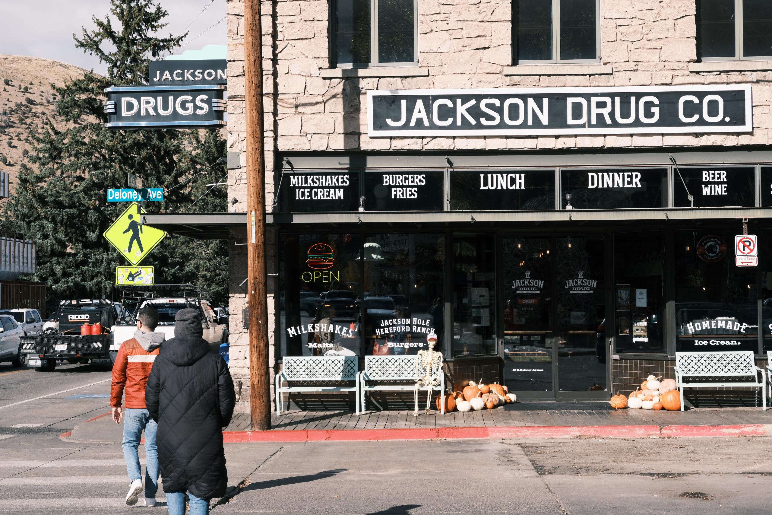 The Jackson Drug Company storefront at the corner of Cache and Deloney in Jackson, Wyoming. There are white benches out front and clusters of Halloween pumpkins decorating the doorway. The windows advertise ice cream, meals, and alcohol for purchase.
