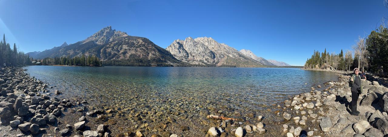 A paraoramic photo of a clear, blue lake. Immediately behind it are tree-covered mountains. On the far right of the frame, a person makes a peace sign and sticks out their tongue at the camera.
