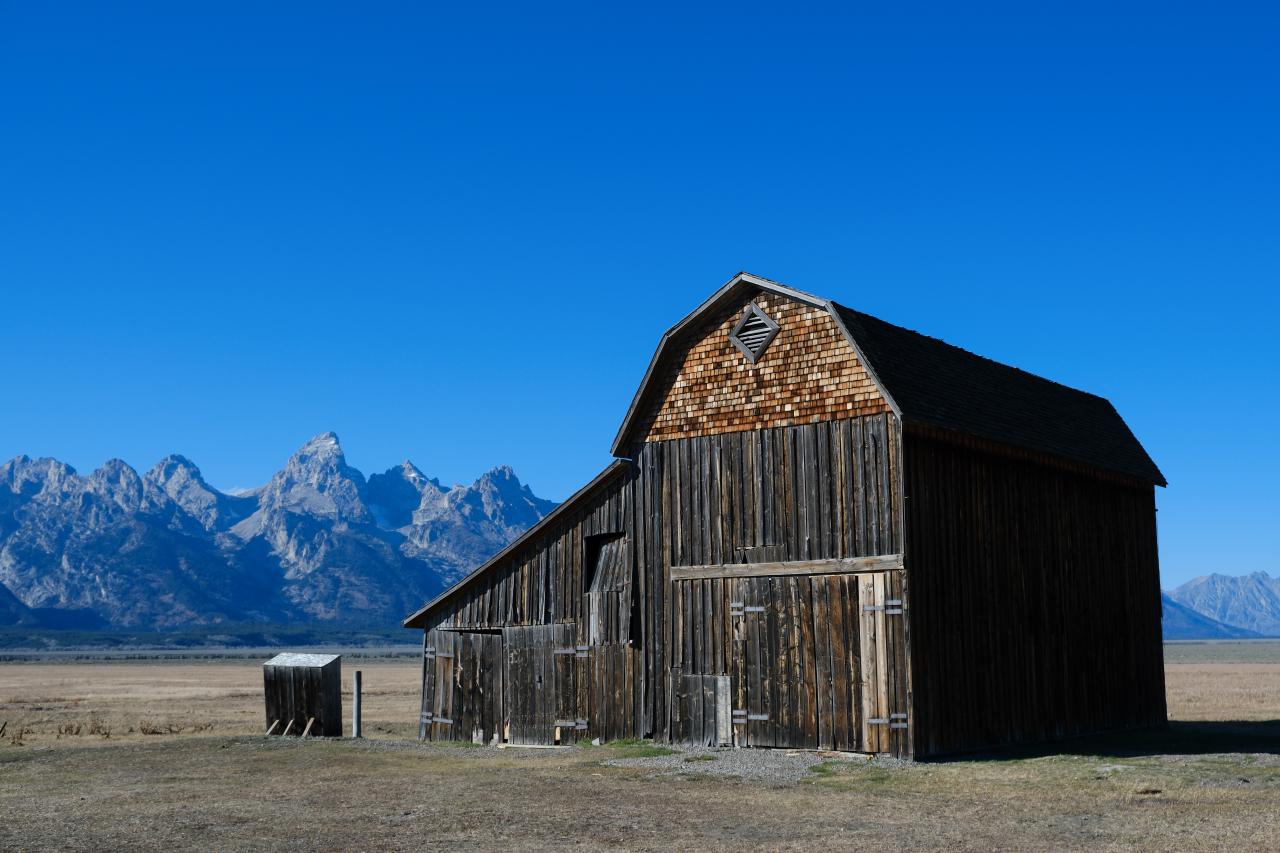 A two-story barn with a variety of siding on each level. The roof of the main part is a traditional barn shape. On the left is a small trapezoidal extension. The Teton mountains are visible in the background.
