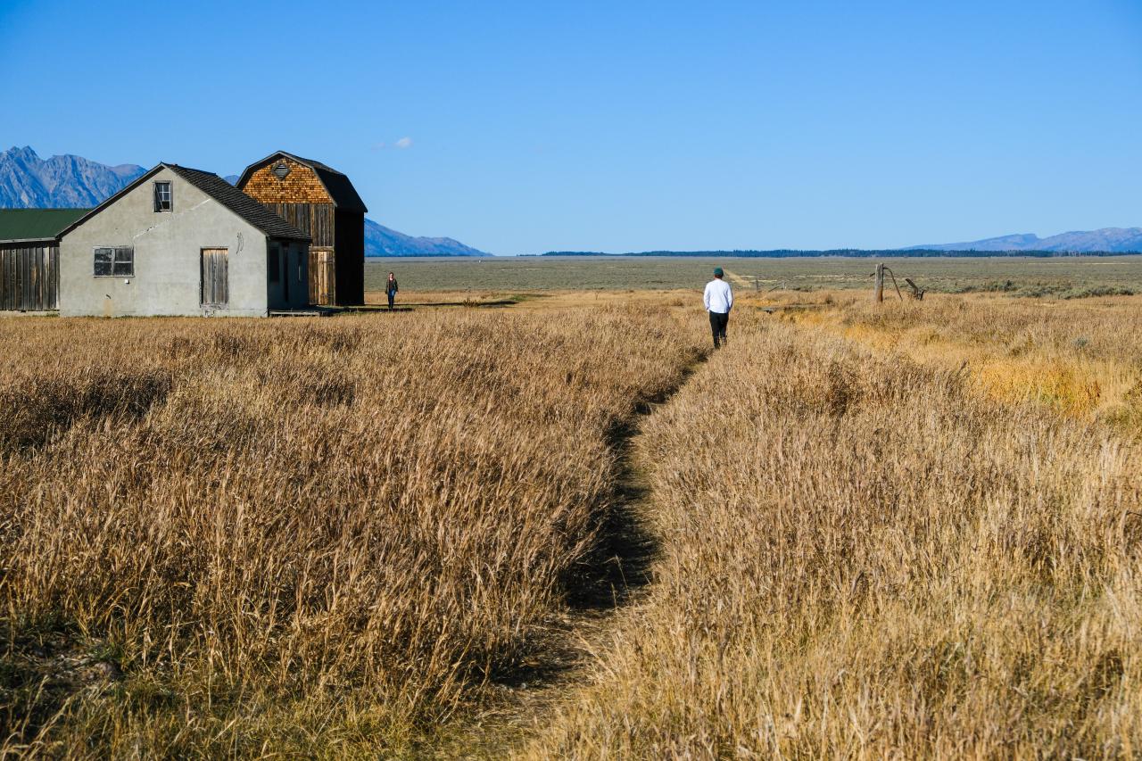 In a dead field of short grasses a small path cuts through. Farther along the path a person wearing a white shirt follows the cut. In the background are a few buildings and one person.
