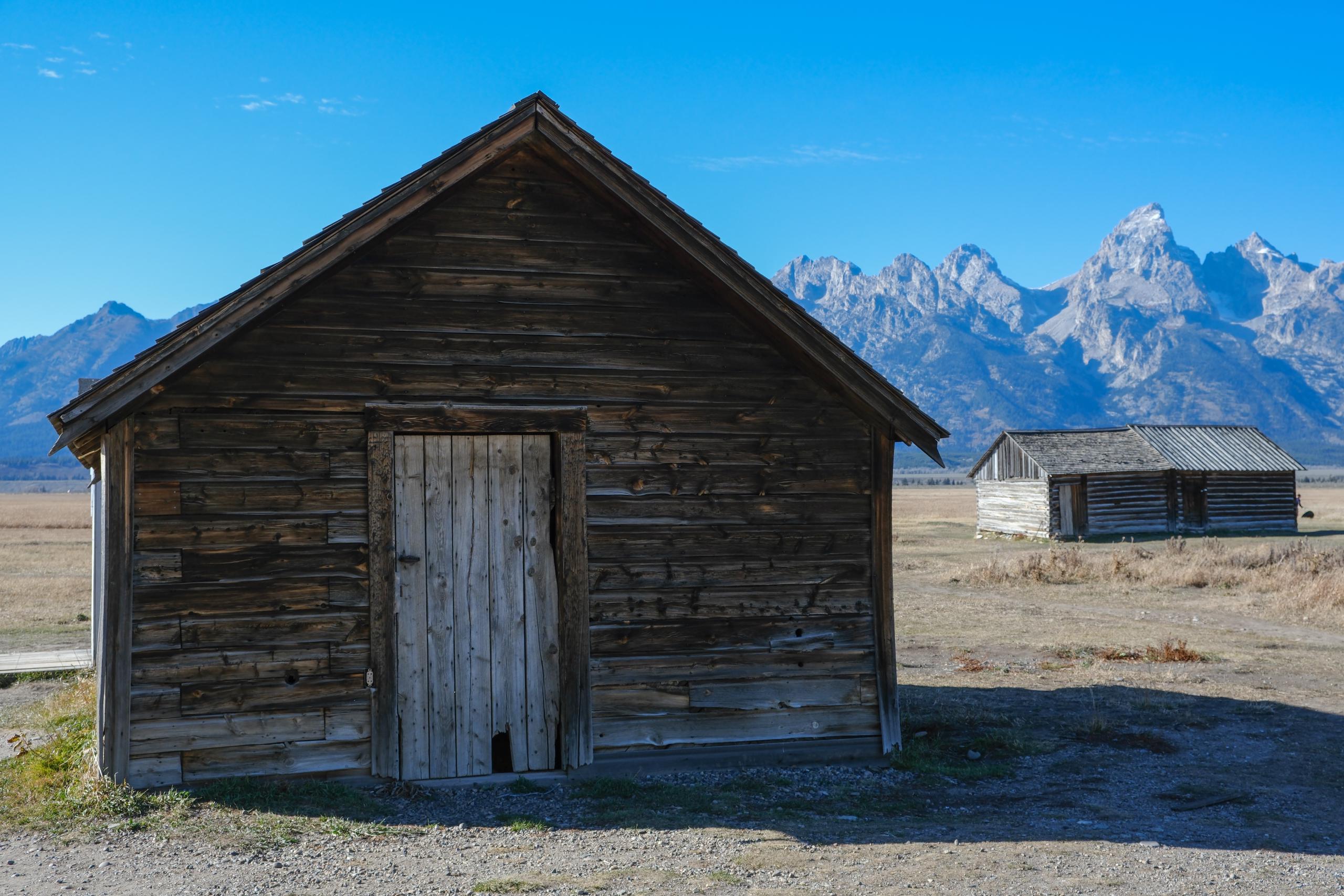 Two weathered cabin-style buildings. The Teton mountains are in the background.
