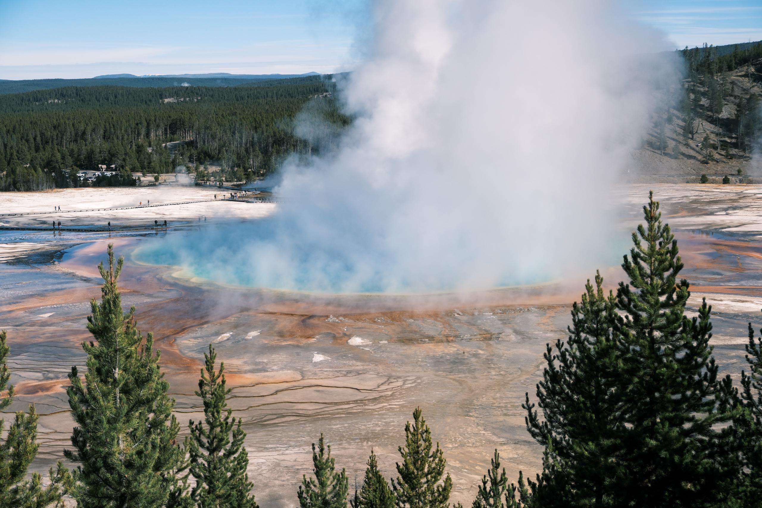 A large hot spring surrounded by flat barren fields. Around the rim are dramatic orange, blue, and yellow rings. Steam billows from the surface.
