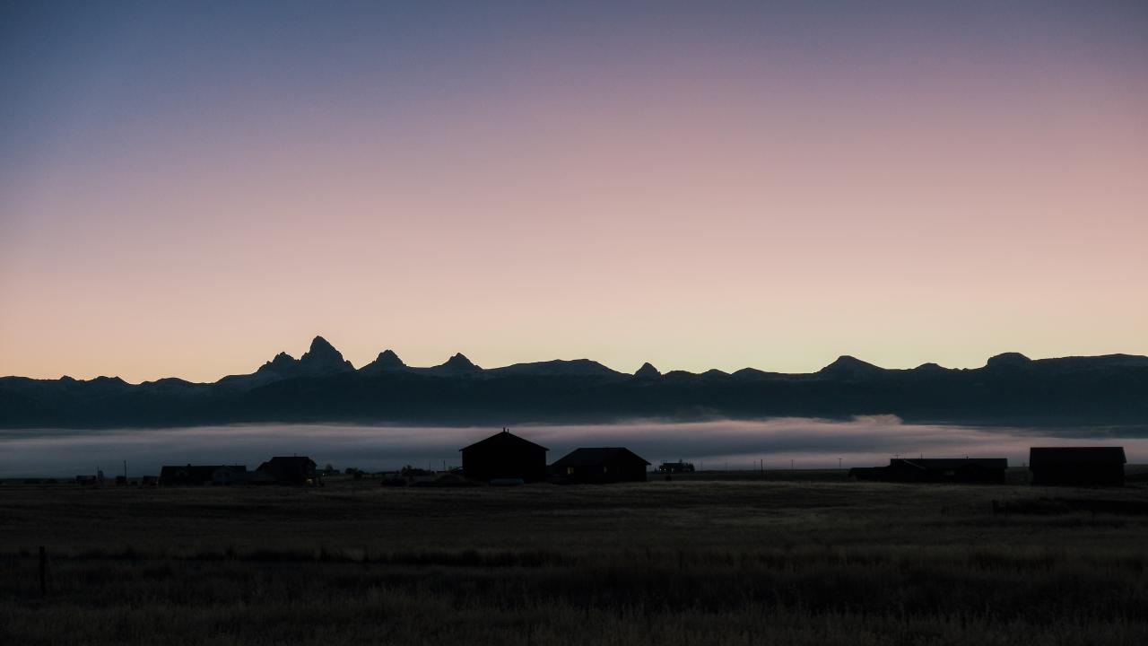 The Teton mountains lit from behind by the rising sun. The sky is colored dramatically in blues, pinks, and purples. In front of the mountains is a mist layer and sillouettes of several farm buildings.
