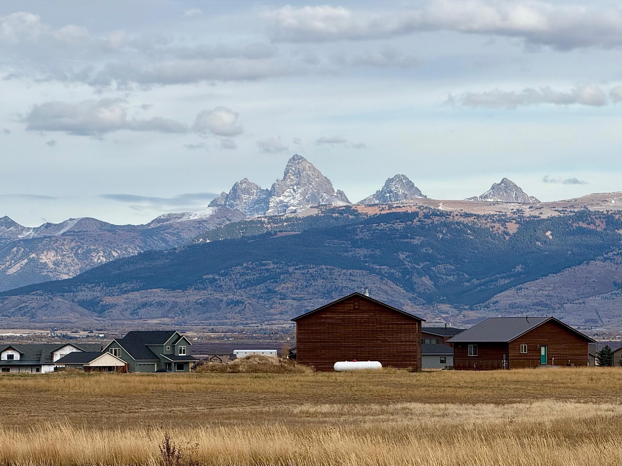 The four Teton peaks appear dramatically above a cluster of buildings in the middle of a field.
