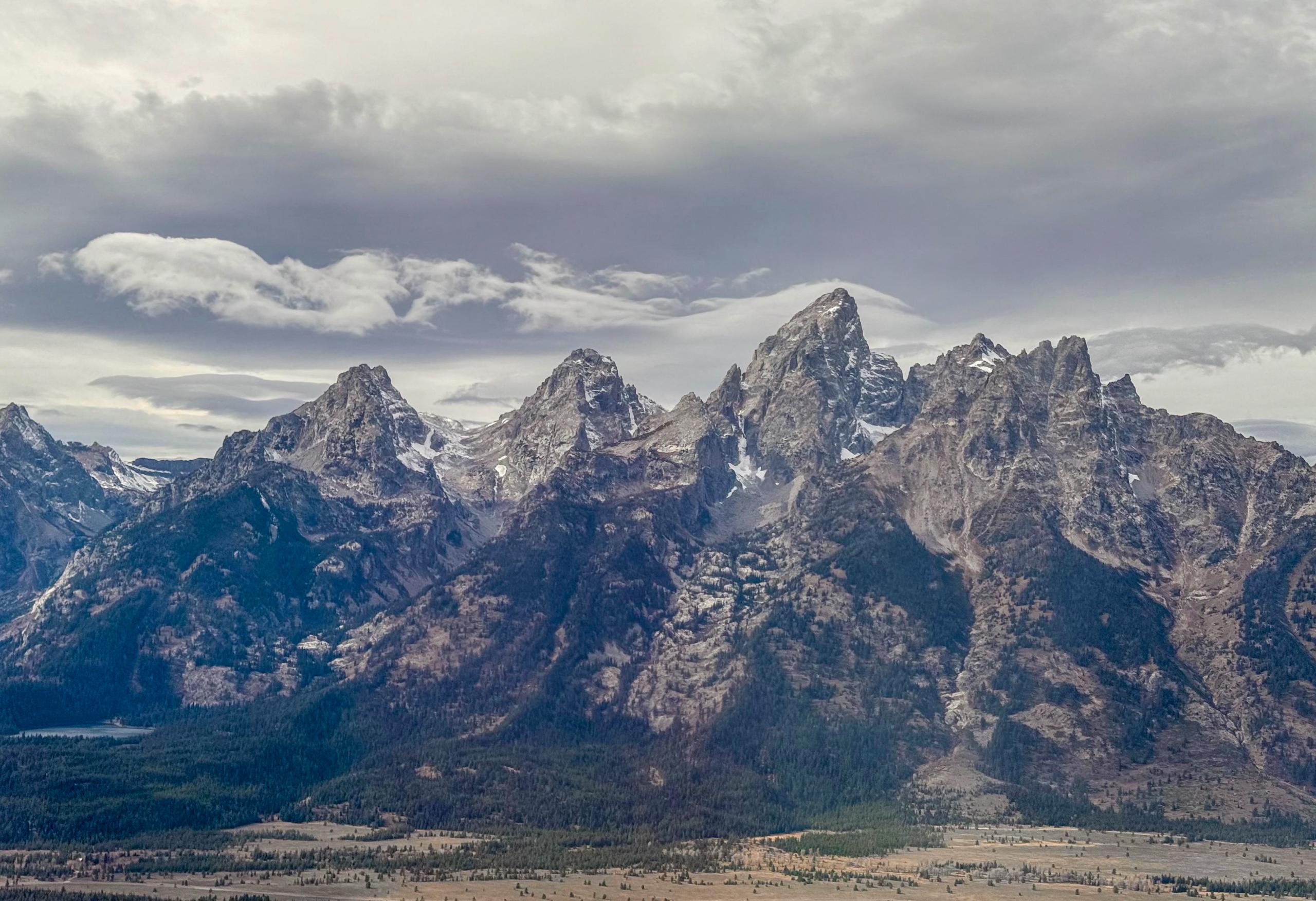 Four craggy peaks rise into a low cloud layer. A streak of flow streaks off of the tallest peak like smoke from a candle. The foothills around these mountains are covered in trees.
