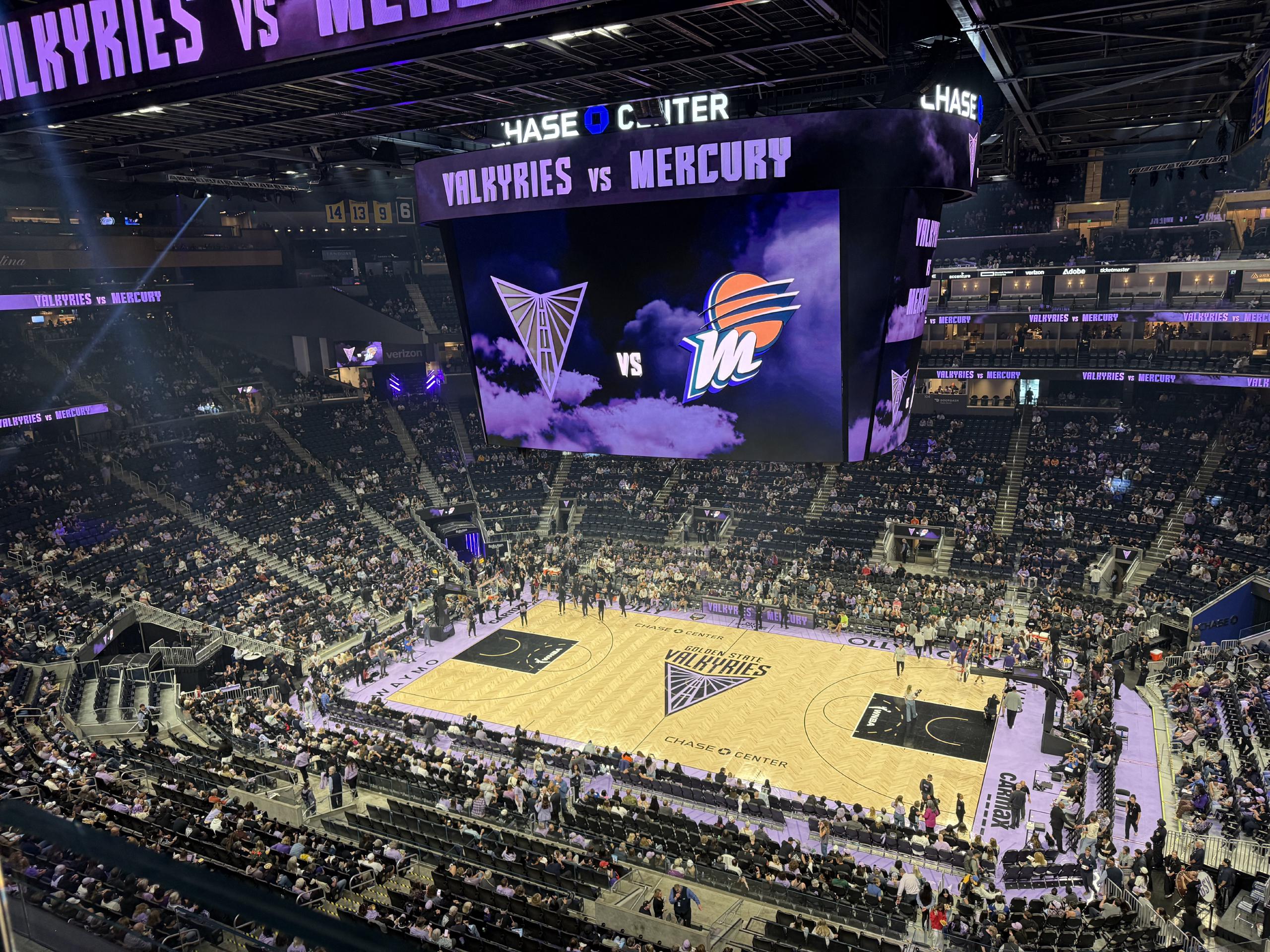 A wide angle view of the court and surrounding stands at Chase Center. The stands are about half full. The large overhead display above the court shows the logos of the two teams. 
