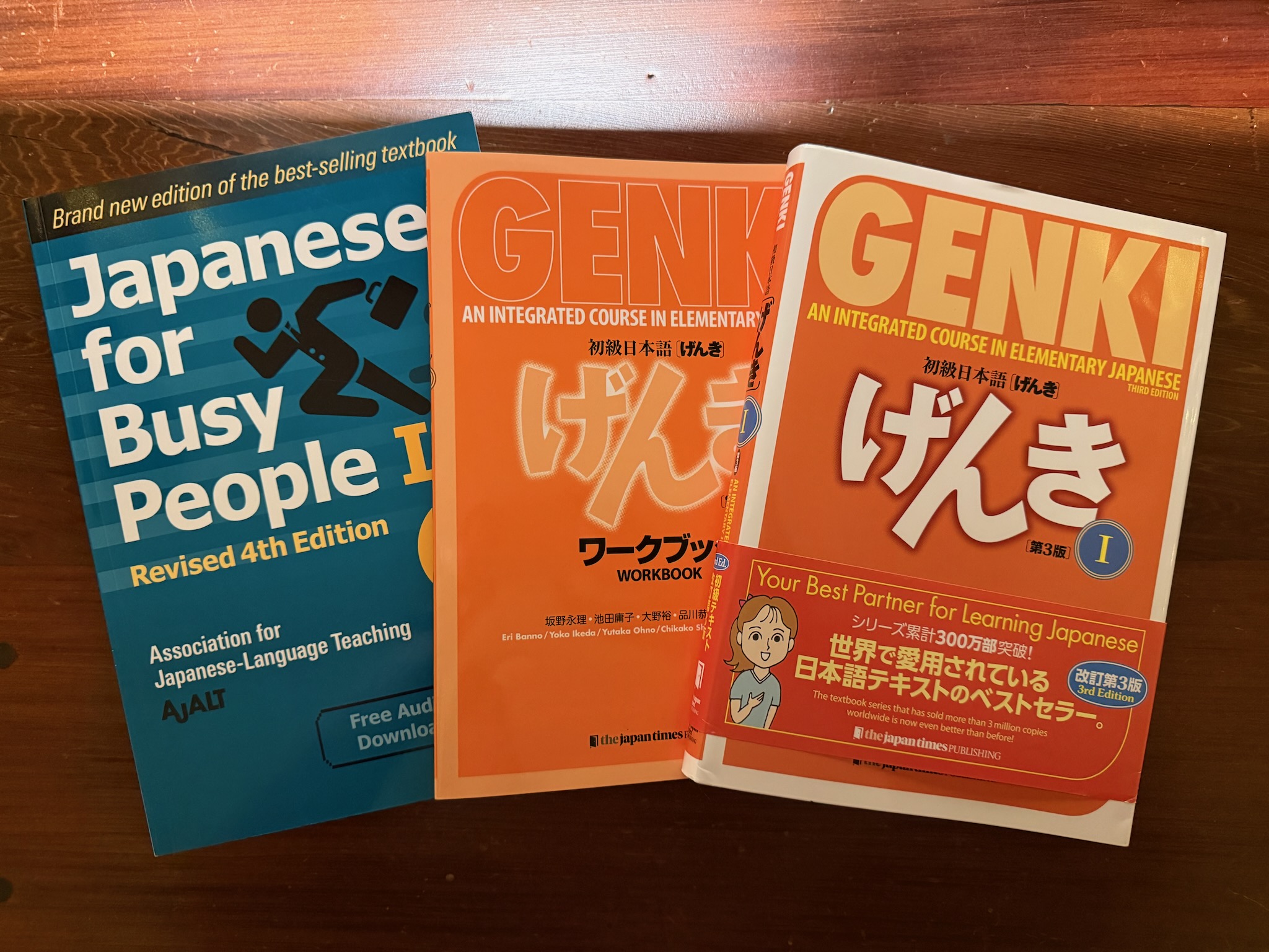 Three textbooks fanned out on a dark wood desktop. From left to right: Japanese for Busy People Book 1, Genki Book 1 Workbook, and Genki Book 1 Textbook.
