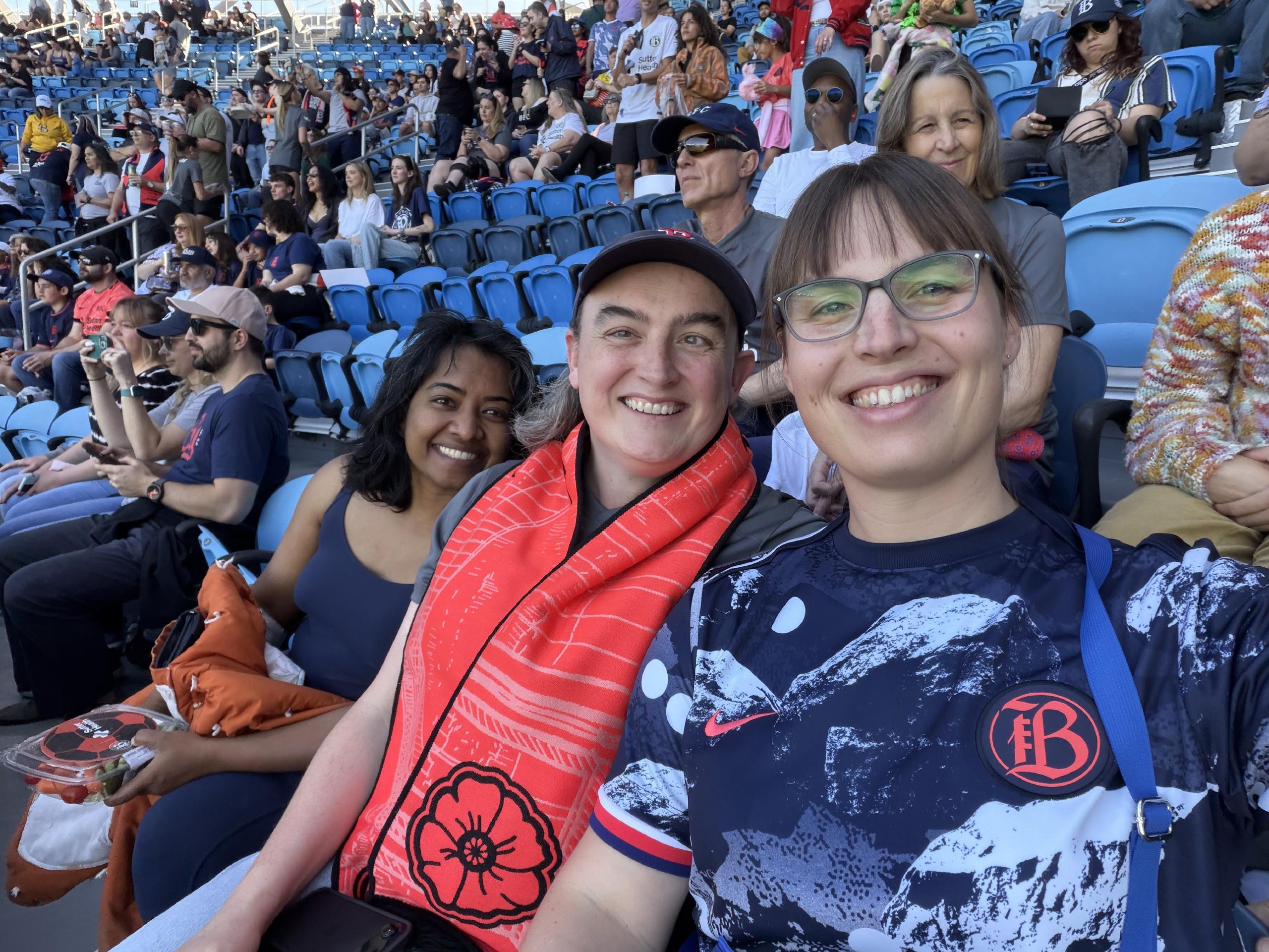 A selfie of three people sitting in stadium seats smiling at the camera.