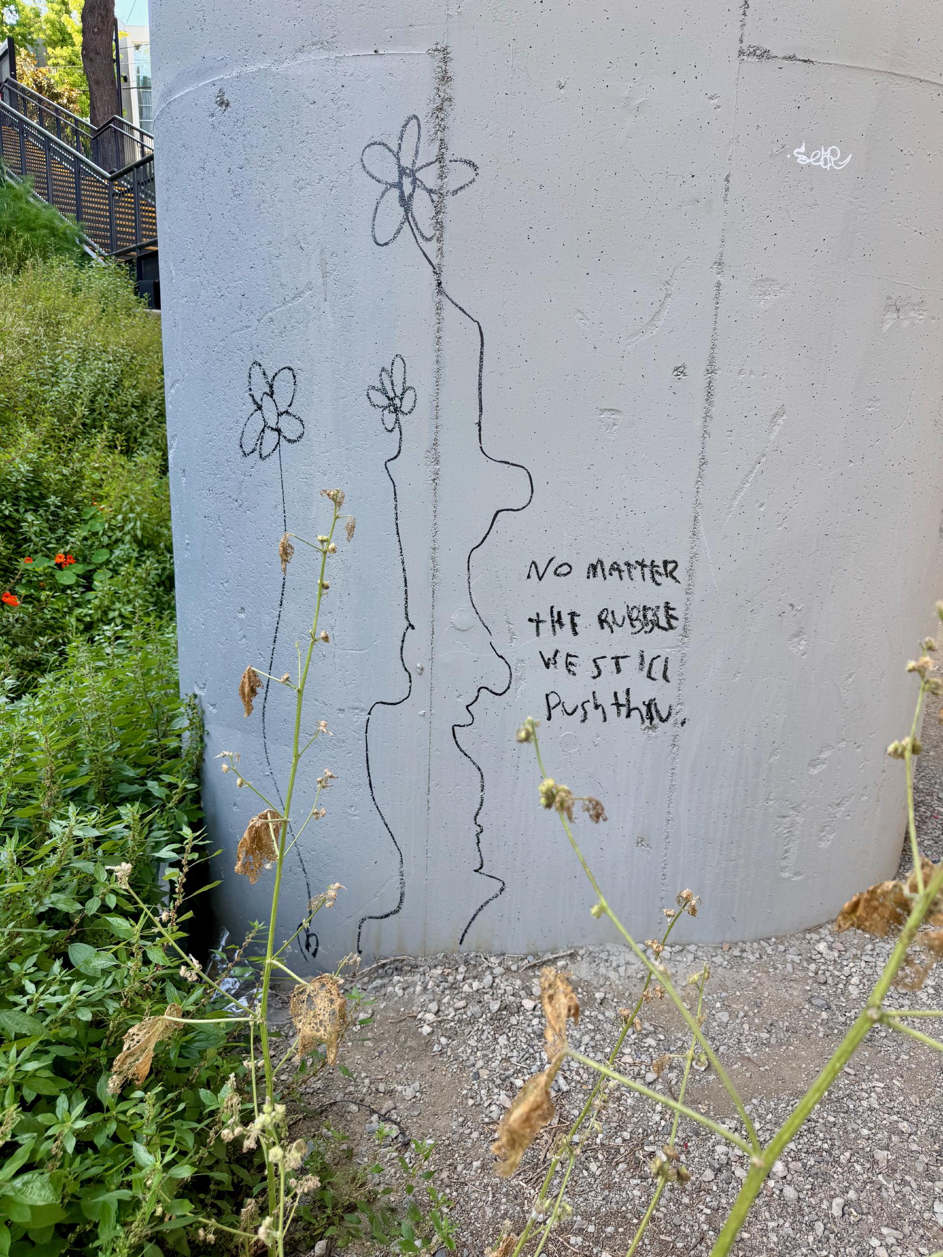 A message scribbled on a concrete pillar says "no matter the rubble we still push thru". There are a few scraggly plants drawn around the text.
