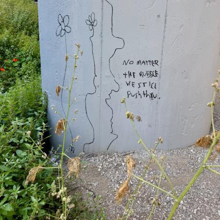 A message scribbled on a concrete pillar says "no matter the rubble we still push thru". There are a few scraggly plants drawn around the text.
