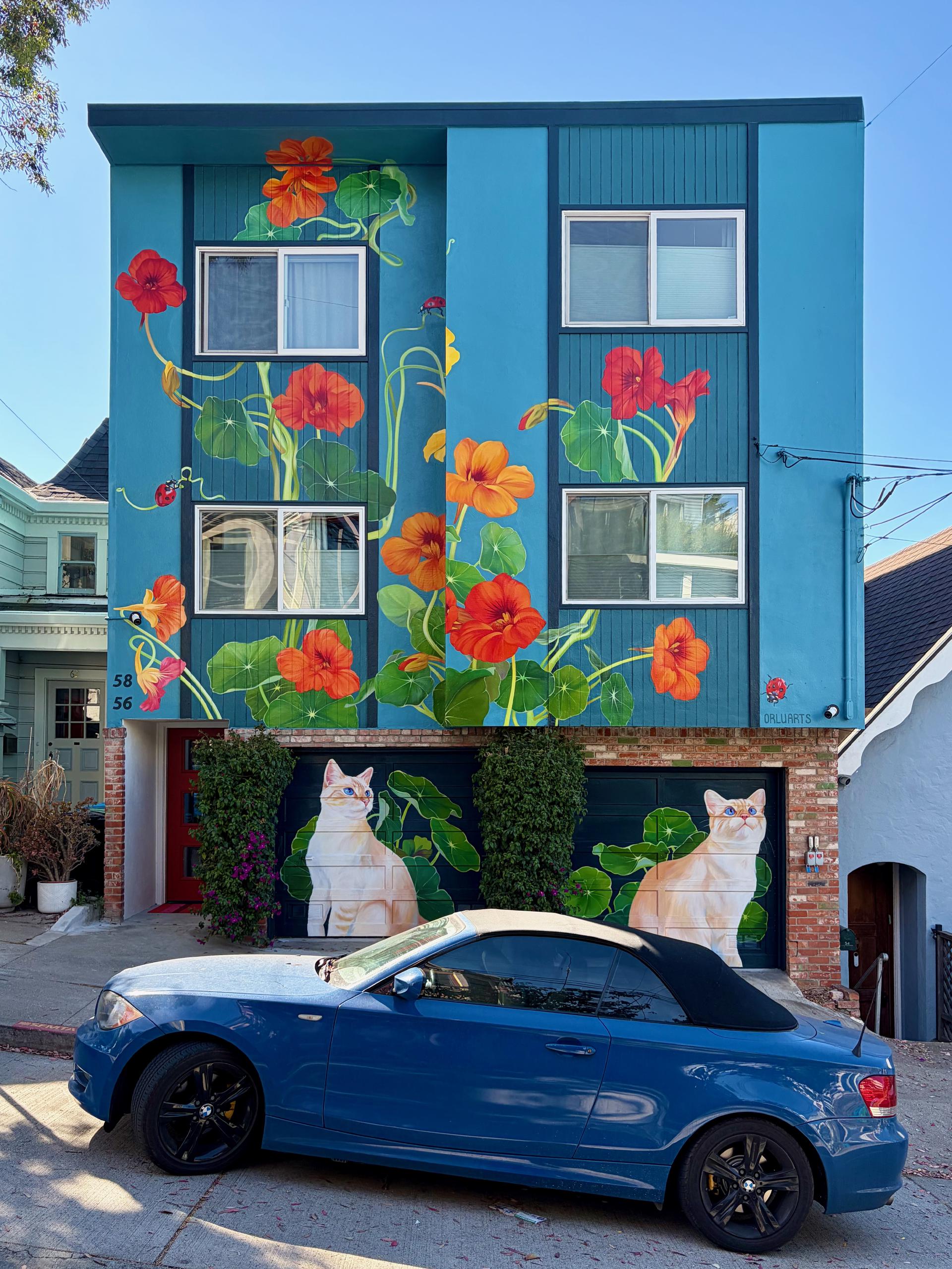 A two-storey house pained various shades of blue. Large California poppies cover the upper stories. The garage at ground level has two cats. There is a blue car parked out front.