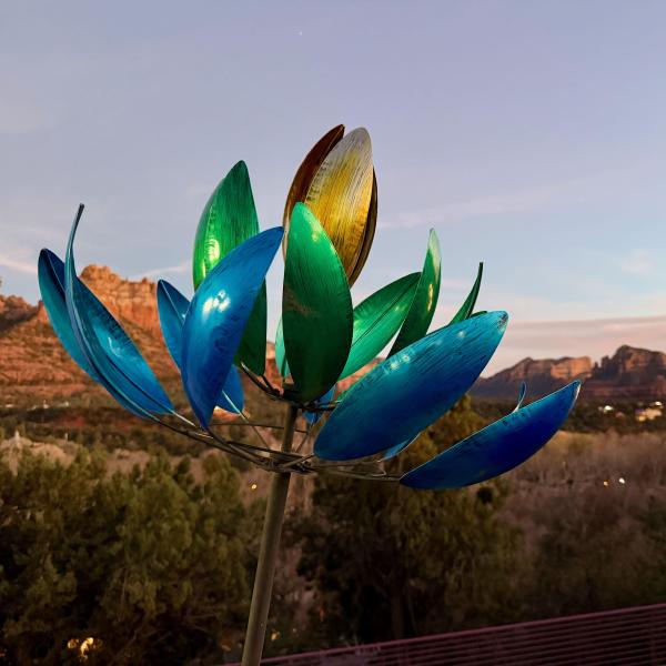 From the bottom left corner, a slender metal stem progresses diagonally toward the center of the image. It erupts into a set of metal petals of varying colors. In the background are the red rocks of Sedona, AZ bathed in evening sunlight.
