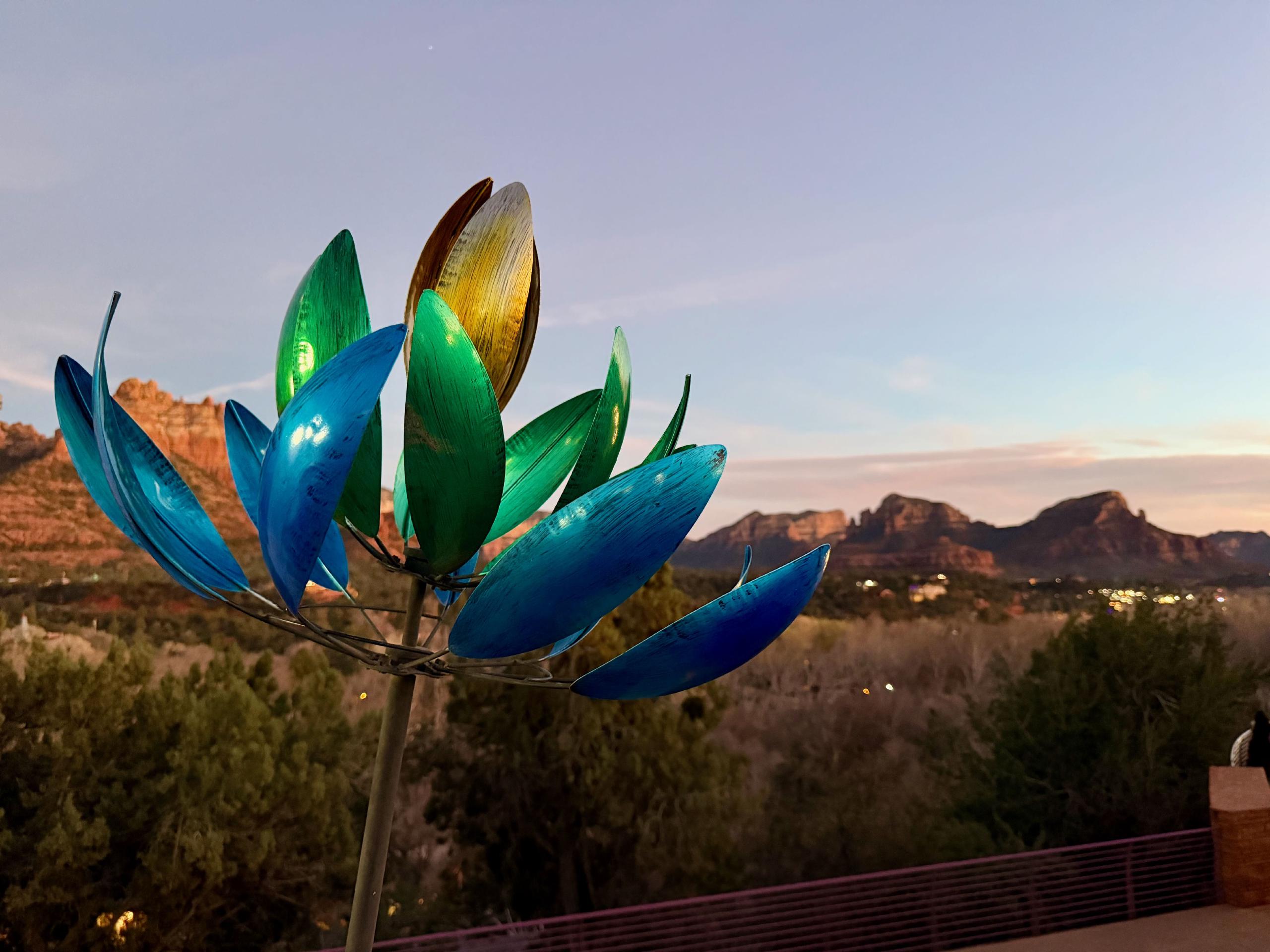 From the bottom left corner, a slender metal stem progresses diagonally toward the center of the image. It erupts into a set of metal petals of varying colors. In the background are the red rocks of Sedona, AZ bathed in evening sunlight.
