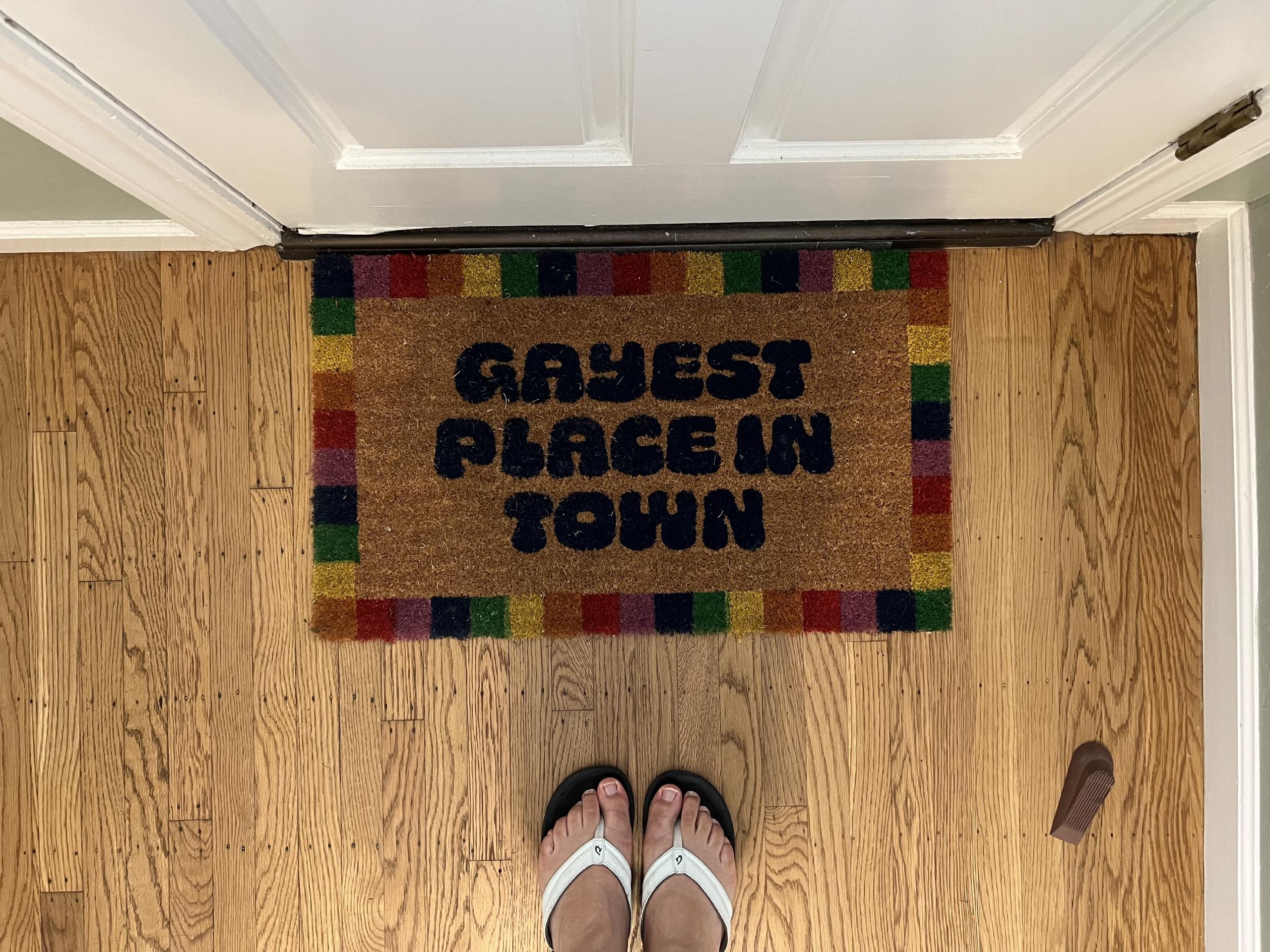 A doormat with a border of rainbow colored blocks. In the middle, in a blobby script, it says 'The Gayest Place In Town'
